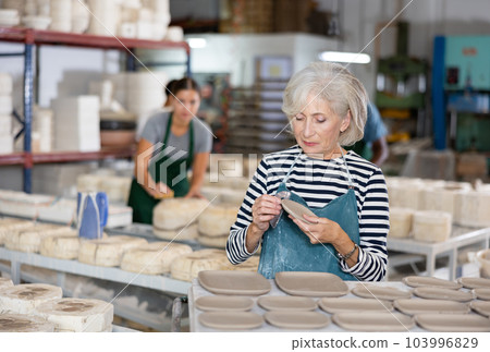 Portrait of elderly woman in apron cutting burrs off freshly made earthenware cups and plates with knife 103996829