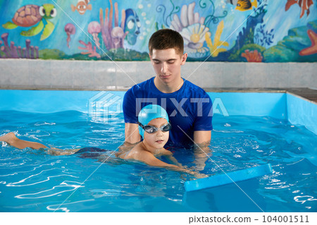 Trainer teaching little boy how to swim in indoor pool with pool floating board 104001511