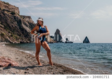 Woman travel sea. Happy tourist in hat enjoy taking picture outdoors for memories. Woman traveler posing on the beach at sea surrounded by volcanic mountains, sharing travel adventure journey 104001711