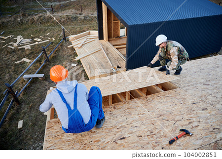 Carpenters mounting wooden OSB board on rooftop of future cottage. Men workers building wooden frame house. Carpentry and construction concept. Carpenters mounting wooden OSB board on rooftop of future cottage. Men workers building wooden frame house. Carpentry and construction concept. 104002851