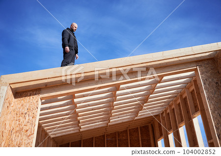 Male designer building wooden frame house. Bald man standing on construction site, inspecting quality of work on sunny day with blue sky on background. Male designer building wooden frame house. Bald man standing on construction site, inspecting quality of work on sunny day with blue sky on background. 104002852