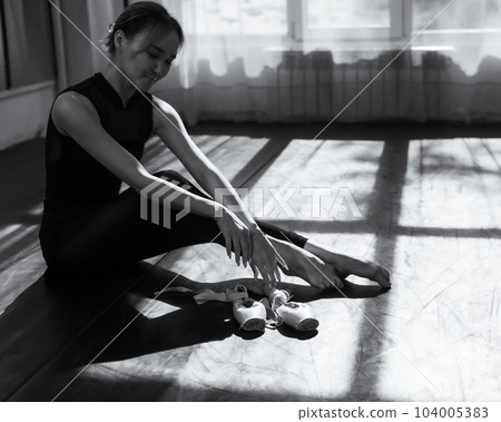 An Asian ballerina sits on the floor and puts on pointe shoes. Monochrome.  104005383