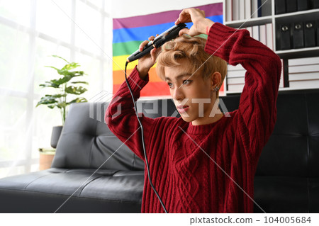 Attractive young gay man in red sweater straightening hair with flat iron. LGBTQ, domestic life, fashion and beauty concept 104005684