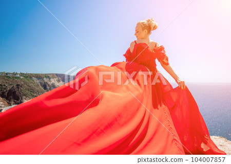 woman sea red dress. Blonde with long hair on a sunny seashore in a red flowing dress, back view, silk fabric waving in the wind. Against the backdrop of the blue sky and mountains on the seashore. 104007857