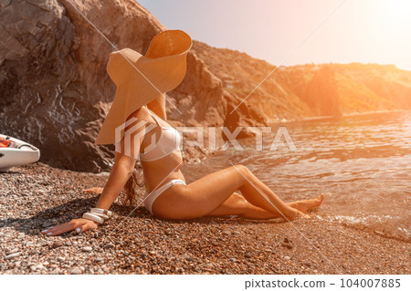 woman sea travel Tanned middle-aged woman with long hair and a white bathing suit. He sits on the seashore in a large sun hat with his back and looks at the sea. 104007885