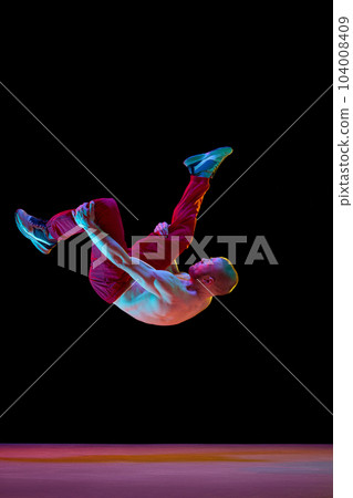 Somersault pose. Artistic young guy with strong muscular body dancing breakdance against black studio background in neon light Somersault pose. Artistic young guy with strong muscular body dancing breakdance against black studio background in neon light 104008409
