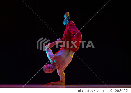Muscular, strong, shirtless young man dancing breakdance, hip hop against black studio background in neon light Muscular, strong, shirtless young man dancing breakdance, hip hop against black studio background in neon light 104008422