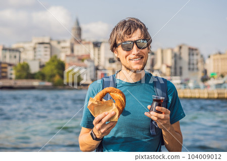 Man in Istanbul having breakfast with Simit and a glass of Turkish tea. Glass of Turkish tea and bagel Simit against golden horn bay and the Galata Tower in Istanbul, Turkey. Turkiye 104009012