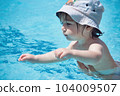 A young white Caucasian toddler boy wearing a hat swimming in a spa pool being supported by his parent 104009507