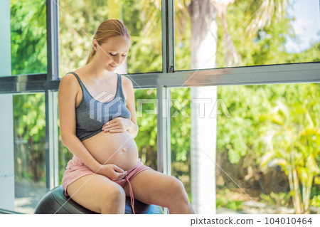 Pregnant woman exercising on fitball at home. Pregnant woman doing relax exercises with a fitness pilates ball. Against the background of the window Pregnant woman exercising on fitball at home. Pregnant woman doing relax exercises with a fitness pilates ball. Against the background of the window 104010464