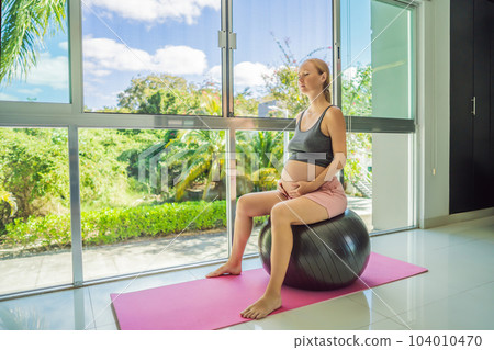 Pregnant woman exercising on fitball at home. Pregnant woman doing relax exercises with a fitness pilates ball. Against the background of the window 104010470