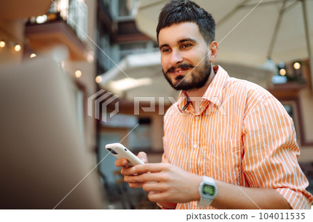 Smiling young man working in cafe on street with laptop and phone. Freelance business concept. Smiling young man working in cafe on street with laptop and phone. Freelance business concept. 104011535