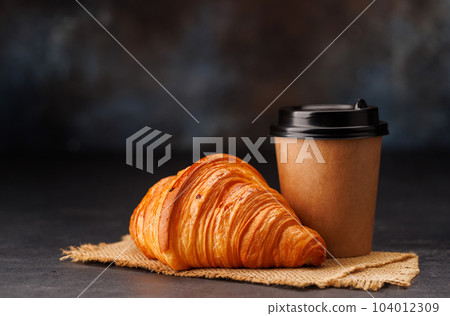 Aromatic coffee in a paper cup paired with a flaky croissant Aromatic coffee in a paper cup paired with a flaky croissant 104012309