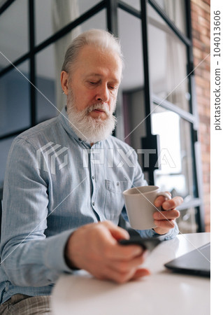 Vertical portrait of elderly mature male with grey hair working on laptop computer and using smartphone, holding cup of coffee, sitting at table in kitchen room in morning, looking device screen. Vertical portrait of elderly mature male with grey hair working on laptop computer and using smartphone, holding cup of coffee, sitting at table in kitchen room in morning, looking device screen. 104013606