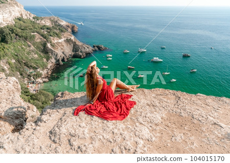 Woman sea red dress yachts. A beautiful woman in a red dress poses on a cliff overlooking the sea on a sunny day. Boats and yachts dot the background. 104015170