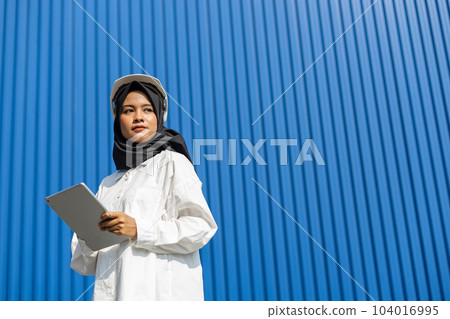 asian muslim woman industrial engineer in uniform wearing safety hard hat using tablet. 104016995