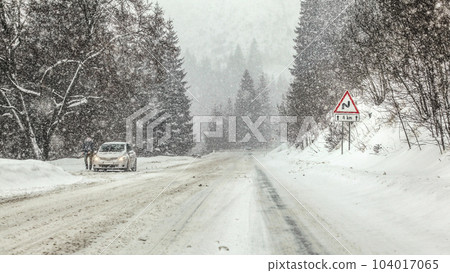 Driving through heavy snowstorm blizzard at forest road, warning curves ahead sign right, and anonymous man with his car stopped on left side of road. View from vehicle driving behind 104017065
