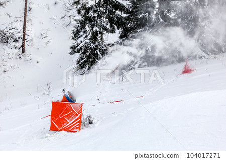 Snow making cannon spraying artificial ice crystals over ski piste, trees in background Snow making cannon spraying artificial ice crystals over ski piste, trees in background 104017271