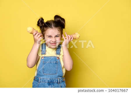 Mischievous naughty Caucasian little kid girl 6 years old in casual denim and yellow t-shirt, making faces, grimacing, posing with ice cream waffle cones, yellow backdrop. Children. Childhood concept 104017667
