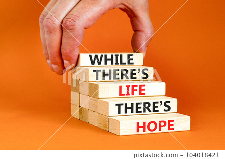 Life and hope symbol. Concept word While there is life there is hope on wooden block. Beautiful orange table orange background. Businessman hand. Business lifestyle life and hope concept. Copy space. 104018421