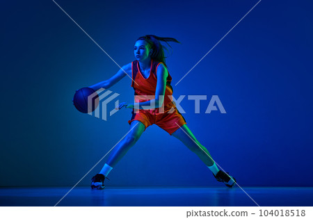 Concentrated female basketball athlete training, dribbling ball against blue studio background in neon light 104018518