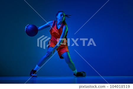 Dynamic image of young girl, basketball player in uniform in motion, playing against blue studio background in neon light 104018519