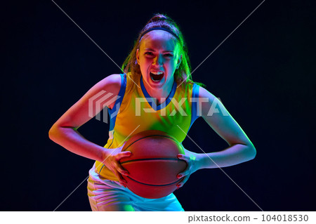Concentrated and motivated young girl, basketball player in uniform posing with ball against black studio background in neon light 104018530
