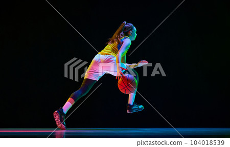 Side bottom view image of young girl, basketball player in motion, running with ball against black studio background in neon light Side bottom view image of young girl, basketball player in motion, running with ball against black studio background in neon light 104018539