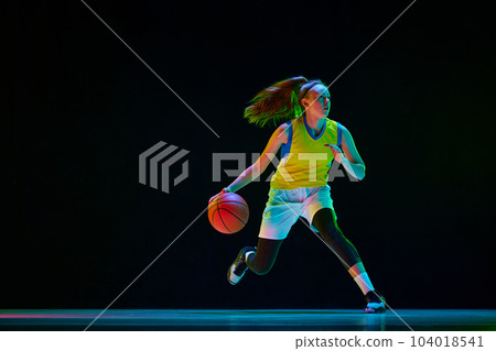 Young motivated girl, basketball player in uniform running with ball against black studio background in neon light 104018541