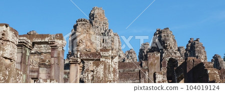 Capture of the ancient Bayon Temple in Cambodia, with its iconic stone towers featuring human faces, on a sunny day. 104019124