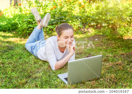 Freelance business concept. Young woman lying on green grass lawn in city park working on laptop pc computer. Lifestyle authentic candid student girl studying outdoors. Mobile Office 104019167
