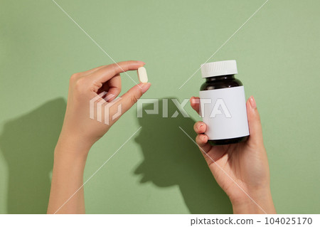 Two hand carrying a small white pill and a blank label medicine bottle. Pastel green background. Mockup of medicine brands 104025170