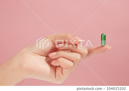 On light pink background, a green capsule standing on a finger of hand model. Medicine and health care concept 104025202