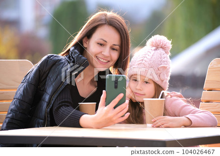 Young mother and her child daughter taking picture with phone selfie camera sitting at street cafe with hot drinks on sunny fall day. Social media presence in everyday life concept 104026467