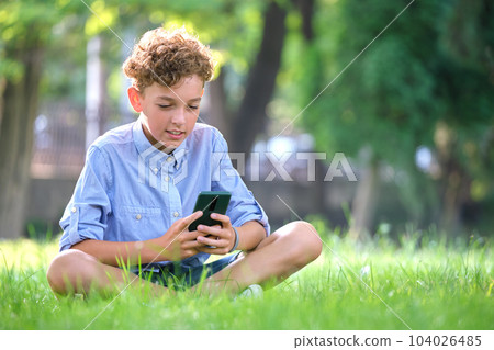 Young boy playing game on his smartphone outdoors in summer park. Addiction from electronic gadgets concept Young boy playing game on his smartphone outdoors in summer park. Addiction from electronic gadgets concept 104026485