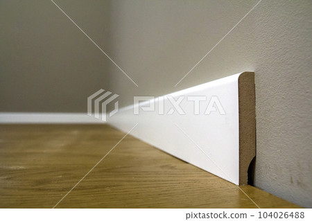 Wooden or plastic white floor plinth installation in big empty room on wooden floor and white plastered stucco walls background. Interior details. 104026488