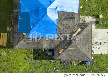 Top view of leaking house roof covered with protective tarp sheets against rain water leaks until replacement of asphalt shingles. Damage of building rooftop as aftermath of hurricane Ian in Florida Top view of leaking house roof covered with protective tarp sheets against rain water leaks until replacement of asphalt shingles. Damage of building rooftop as aftermath of hurricane Ian in Florida 104026777