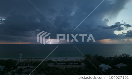 Seashore town with dark houses and tall lighthouse on ocean shore with blinking light at stormy night for vessels navigation. Thunderstorm with lightnings over sea water posing danger for ships 104027101