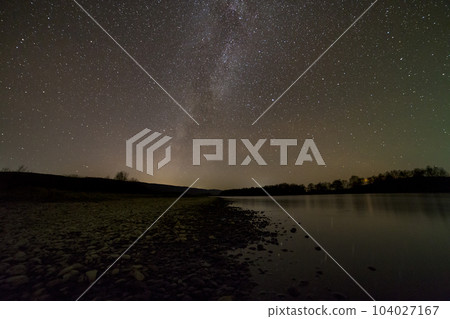 Peaceful landscape panorama at night. Long exposure shot of pebbles river bank, trees on horizon, bright stars and Milky Way galaxy in dark sky reflected in quiet water. Beauty of nature concept. Peaceful landscape panorama at night. Long exposure shot of pebbles river bank, trees on horizon, bright stars and Milky Way galaxy in dark sky reflected in quiet water. Beauty of nature concept. 104027167