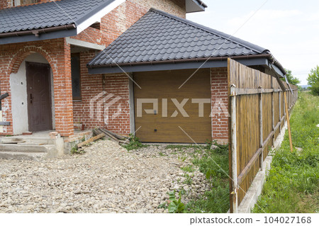 Part view of modern two-storied not finished new brick family cottage house with steep brown shingled roof, attached garage, temporary wooden fence. Construction, investment, real property concept. 104027168