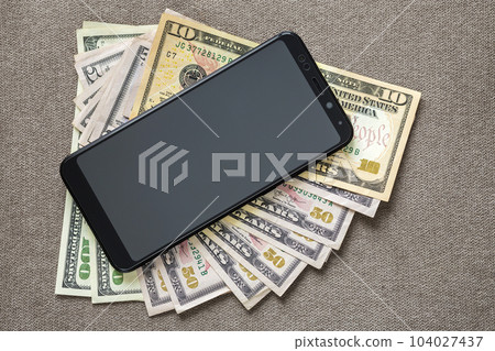 New black modern cellphone on money dollars banknotes background. Modern technology, communication and online trade using gadget concept. New black modern cellphone on money dollars banknotes background. Modern technology, communication and online trade using gadget concept. 104027437
