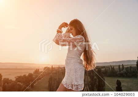 Happy woman in white boho dress on sunset in mountains. Romantic woman with long hair standing with her back on the sunset in nature in summer with open hands. Silhouette. Nature. Sunset. Happy woman in white boho dress on sunset in mountains. Romantic woman with long hair standing with her back on the sunset in nature in summer with open hands. Silhouette. Nature. Sunset. 104029003