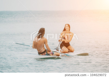 Sea woman and man on sup. Silhouette of happy young woman and man, surfing on SUP board, confident paddling through water surface. Idyllic sunset. Active lifestyle at sea or river. 104029072