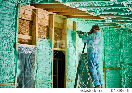 Male builder insulating wooden frame house. Man worker spraying polyurethane foam inside of future cottage, standing on ladder, using plural component gun. Construction and insulation concept. 104030696