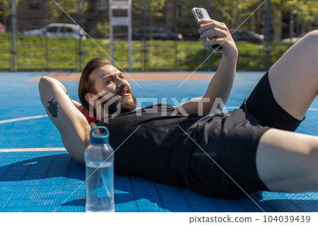 Tired man in sportswear after playing basketball game lying down on ball relaxing using smartphone 104039439