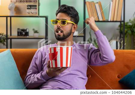 Indian man sits on couch eating popcorn and watching interesting TV serial sport game online at home 104044206