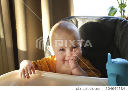 Toddler girl in a yellow T-shirt sits on a highchair and dines on biscuit. Little baby learning to eat on her own. Child eats with hands. Lunch at the baby in high chair. Baby sitting and smiling. 104044584