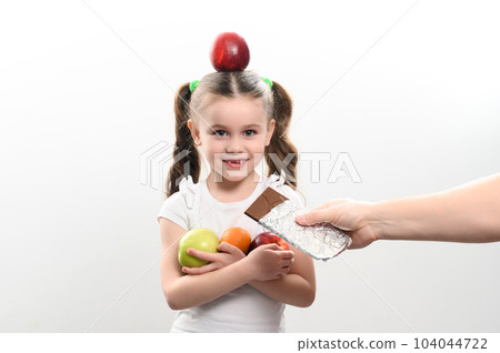 Portrait of a child with an apple on his head and fruit in his hands on a white background, a girl with fruit is offered chocolate. Portrait of a child with an apple on his head and fruit in his hands on a white background, a girl with fruit is offered chocolate. 104044722
