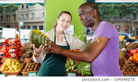 Smiling seller woman is helping man choose melone in the fruit market. 104046078