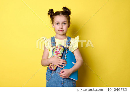 Confident adorable smart primary school girl holding copybook and pencil case, going to school, smiling looking at camera, isolated on yellow background with copy ad space. Back to school concept 104046783